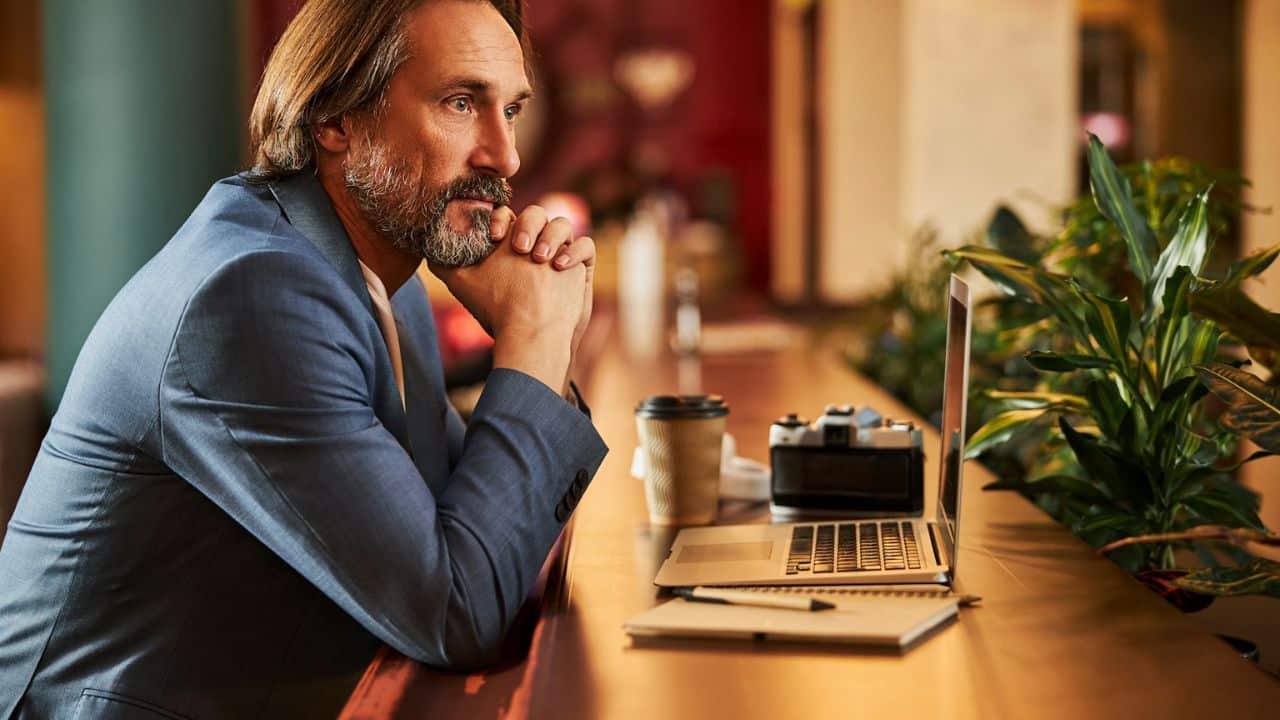 A thoughtful man with a beard is leaning on a counter near a laptop and camera.