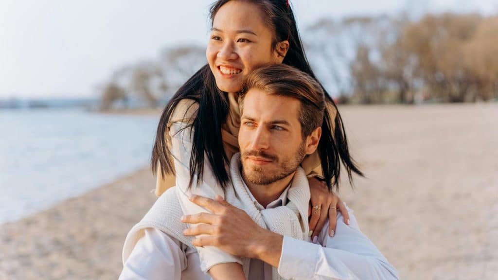 A woman hugging a man from behind as they pose together on a beach.