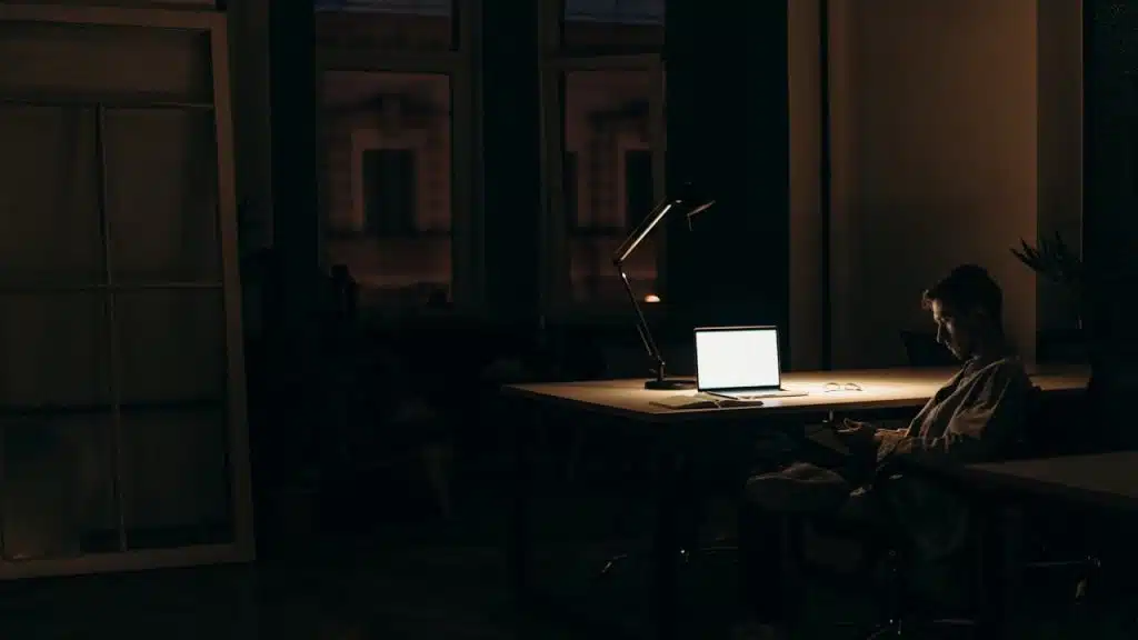 A person is sitting at a desk in a dark room late at night, working on a laptop with a desk lamp providing light.