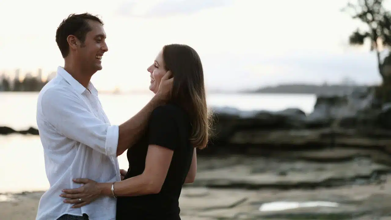 A smiling couple embraces on a beach at sunset, looking into each other's eyes.