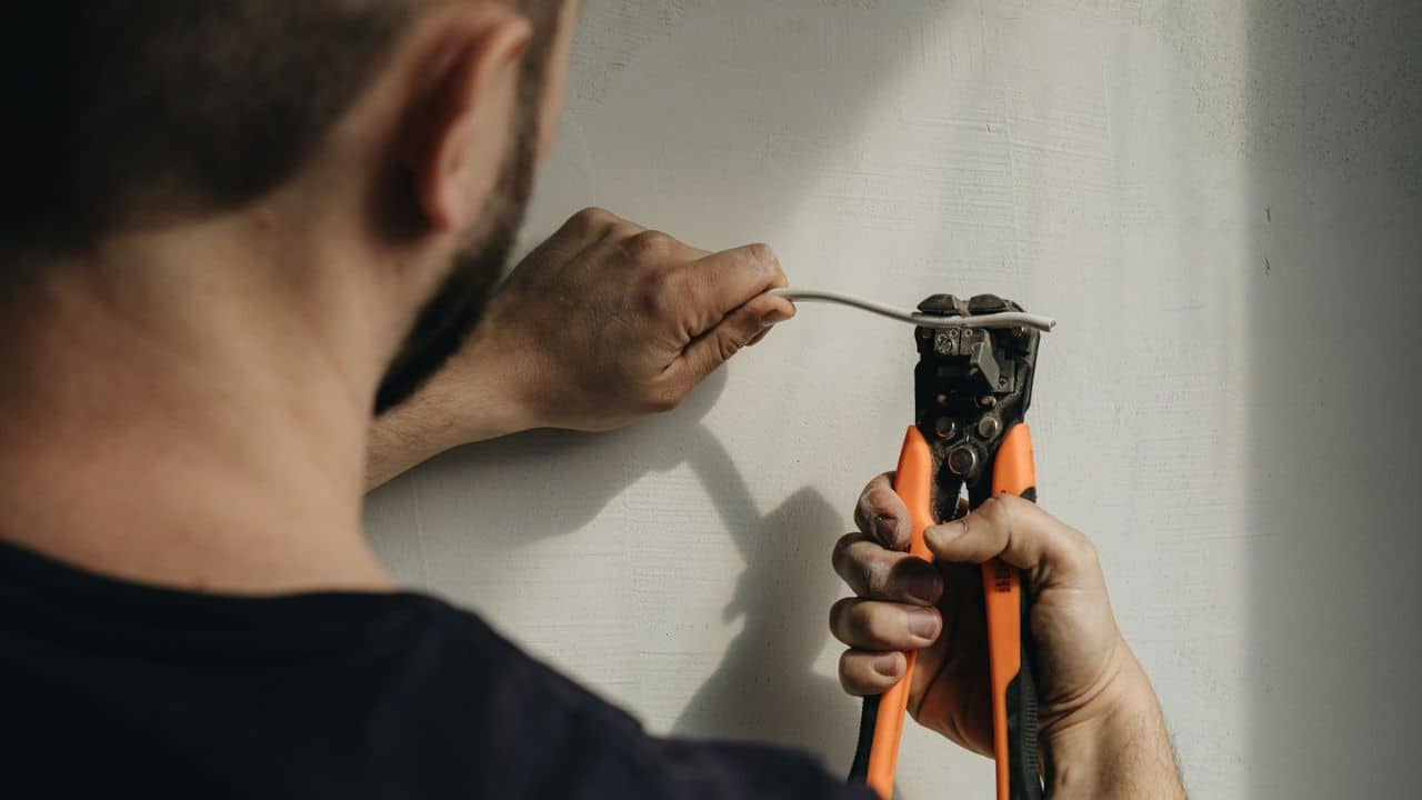 A man is stripping a white wire using orange and black wire strippers against a wall.