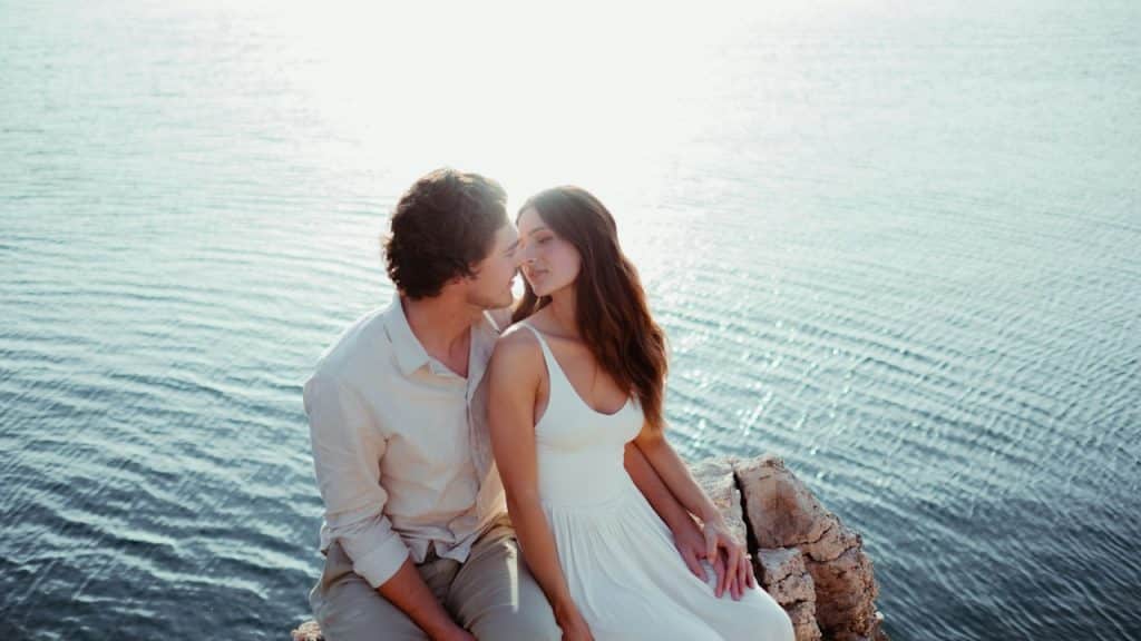 A man and a woman sitting on a rock by the water.