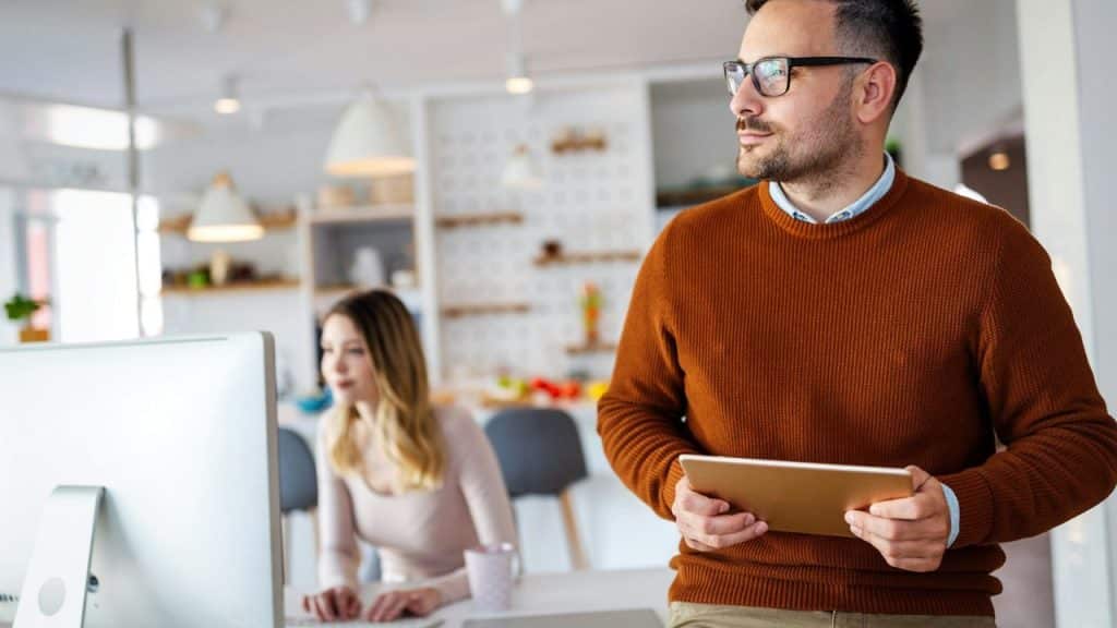 A man in glasses holds a tablet while a woman works on a desktop computer.