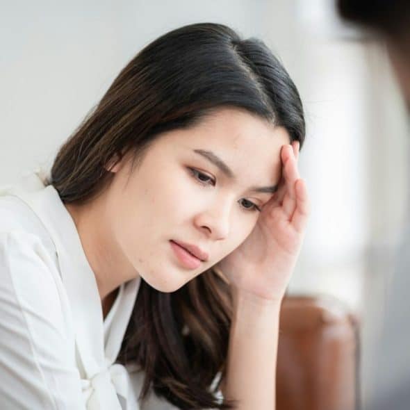 A young woman with long dark hair sits and rests her forehead on her hand.