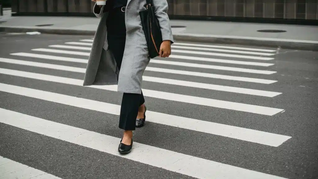 A person in a gray coat walking across a crosswalk with coffee and a bag.