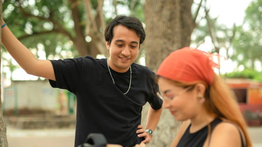 A man is standing and smiling while a woman, wearing an orange bandana, looks down at something she is holding outdoors.