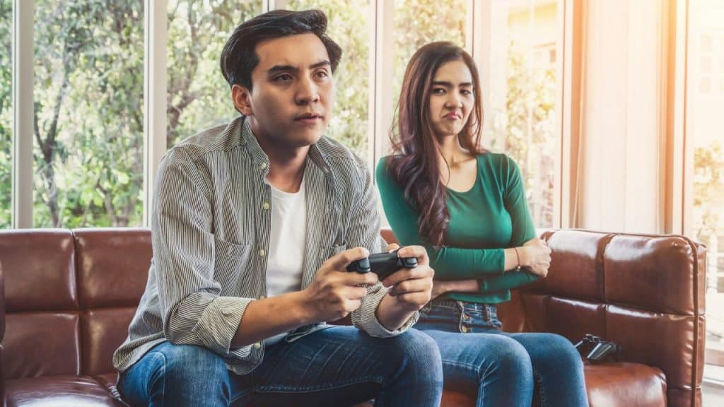 A man plays a video game while a displeased woman sits beside him with crossed arms.