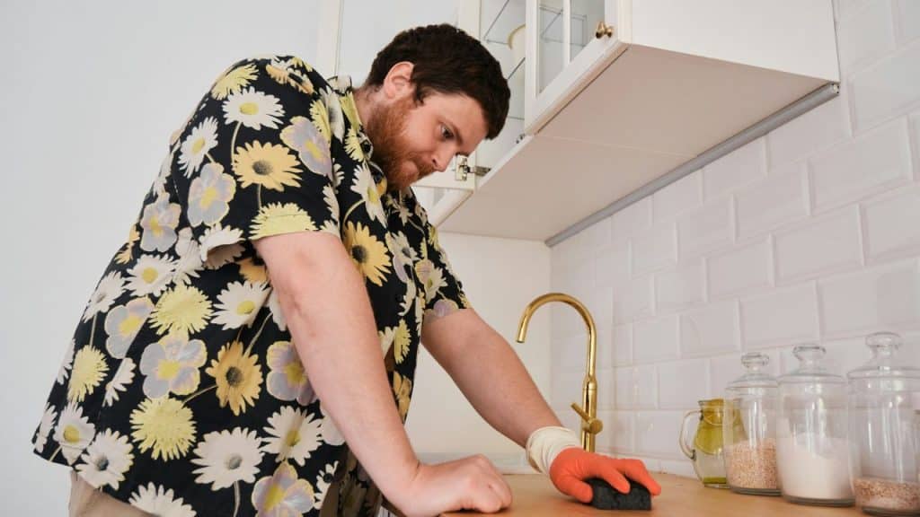 Man in a floral shirt wearing an orange glove leans over a sink and wipes a counter.