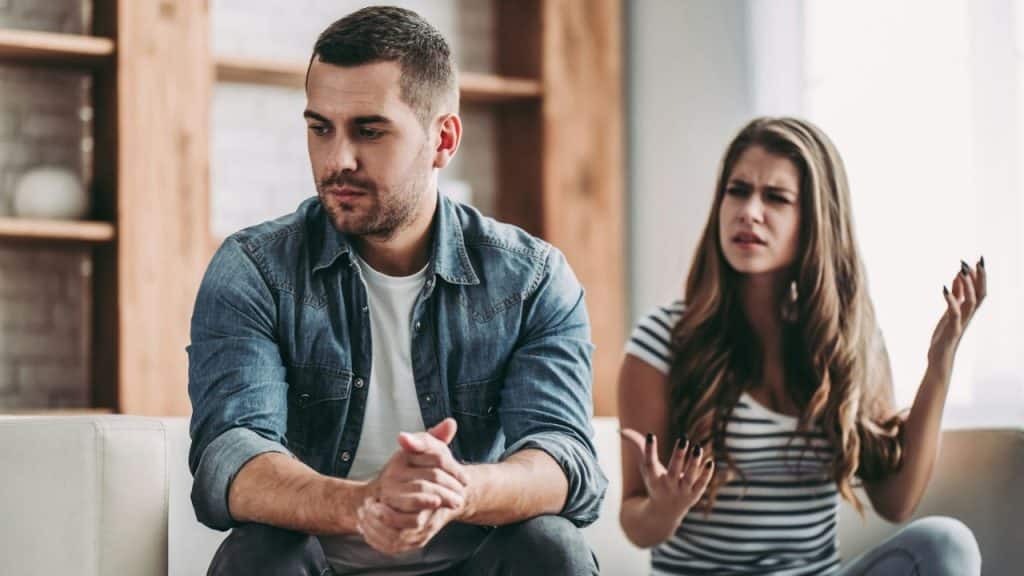 A man sits looking away while a woman argues and gestures with her hands.