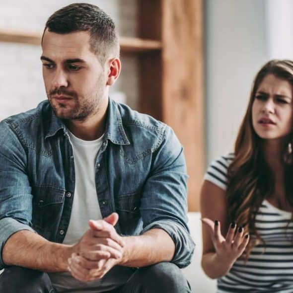 A man sits looking away while a woman argues and gestures with her hands.