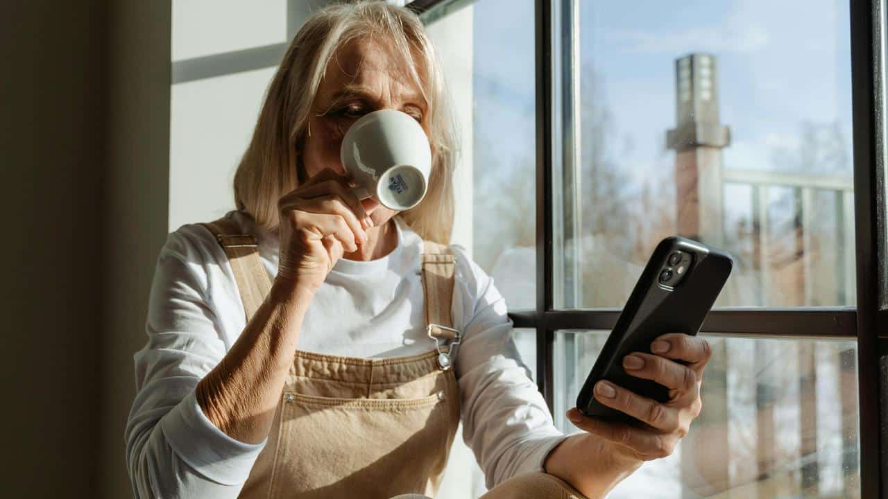 A woman drinking coffee while looking at her phone by a window.