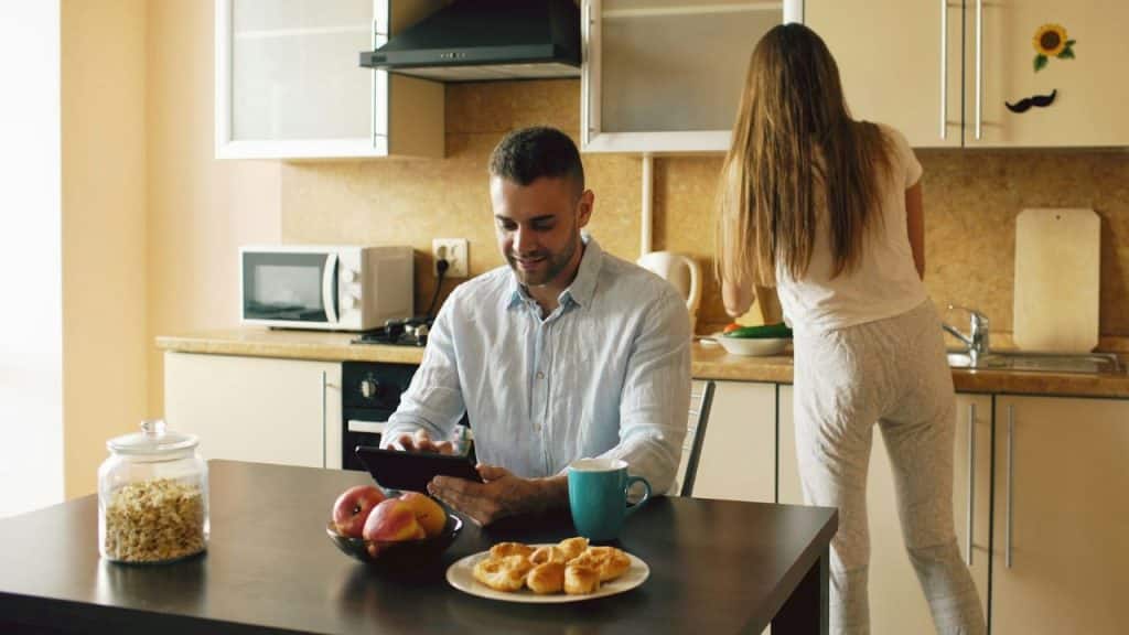 A man uses a tablet at a kitchen table while a woman stands with her back to him.