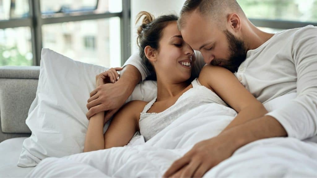 A couple is affectionately embracing in bed under white covers.