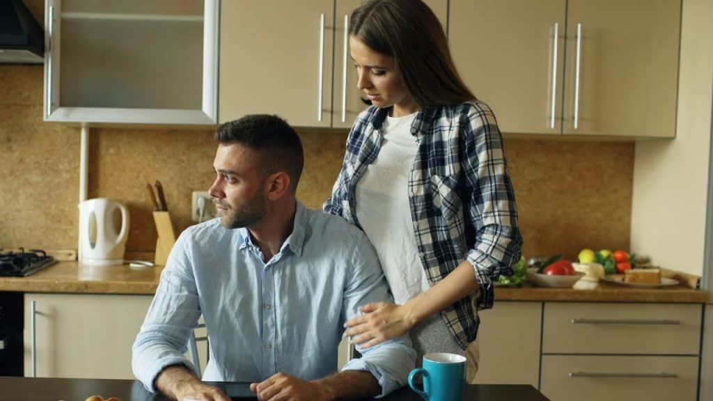 A woman stands behind a man sitting at a kitchen table, placing a hand on his shoulder.
