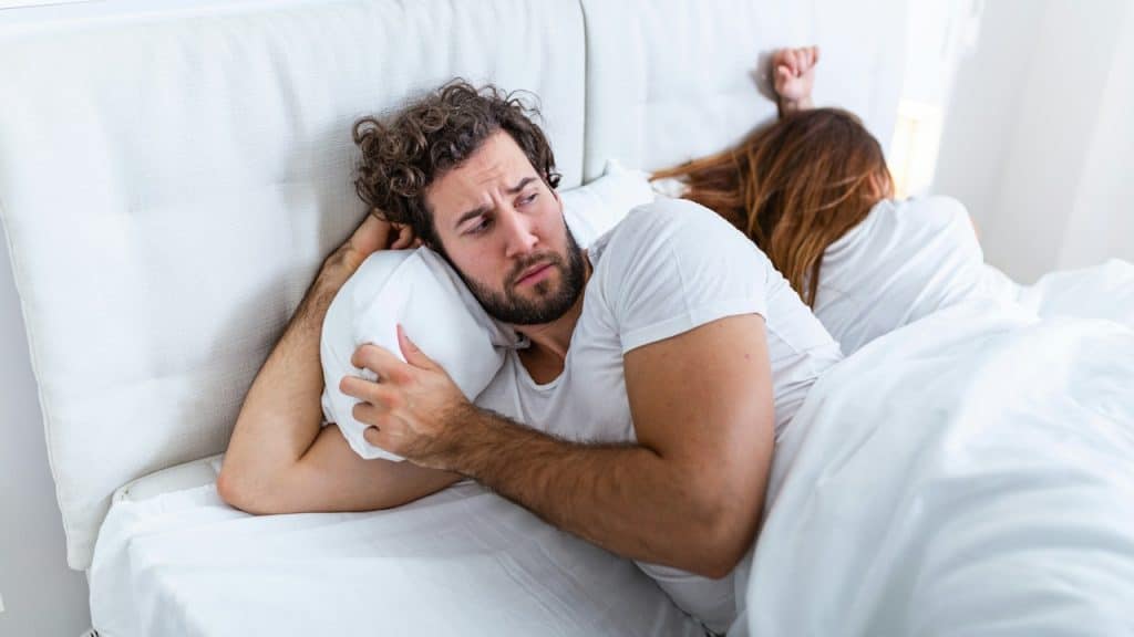 A man is lying in bed looking stressed or anxious, while a woman lies next to him with her back turned.