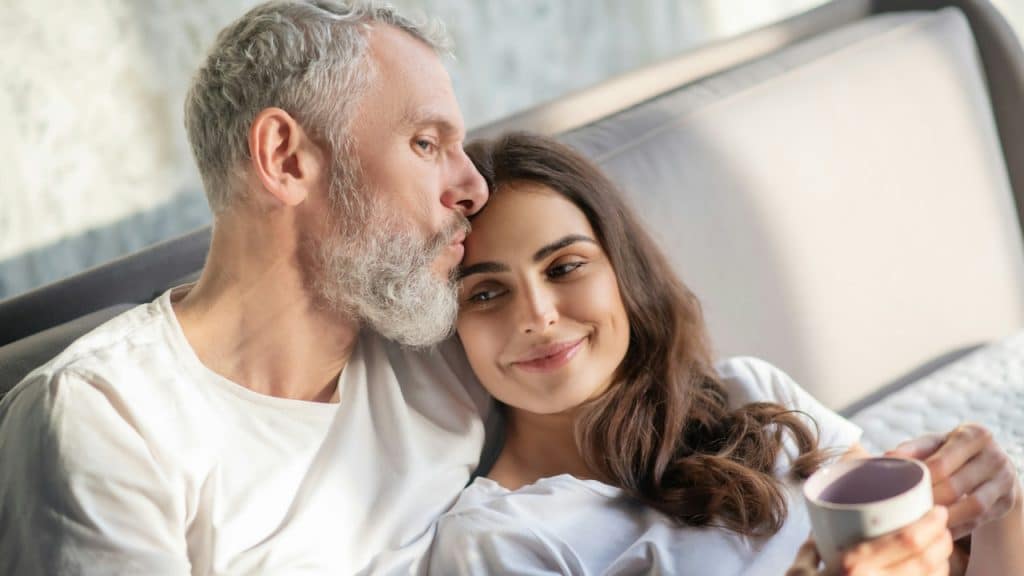An older bearded man kissing a younger woman holding a coffee mug while they recline on a sofa.