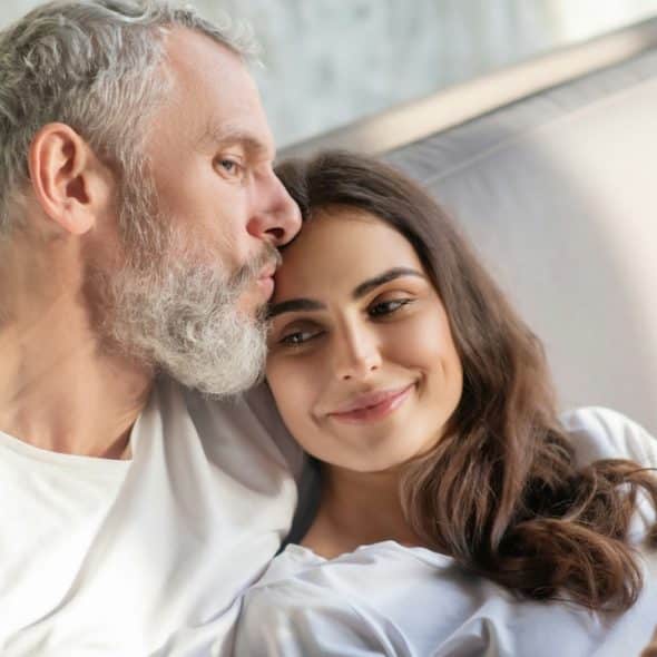 An older bearded man kissing a younger woman holding a coffee mug while they recline on a sofa.