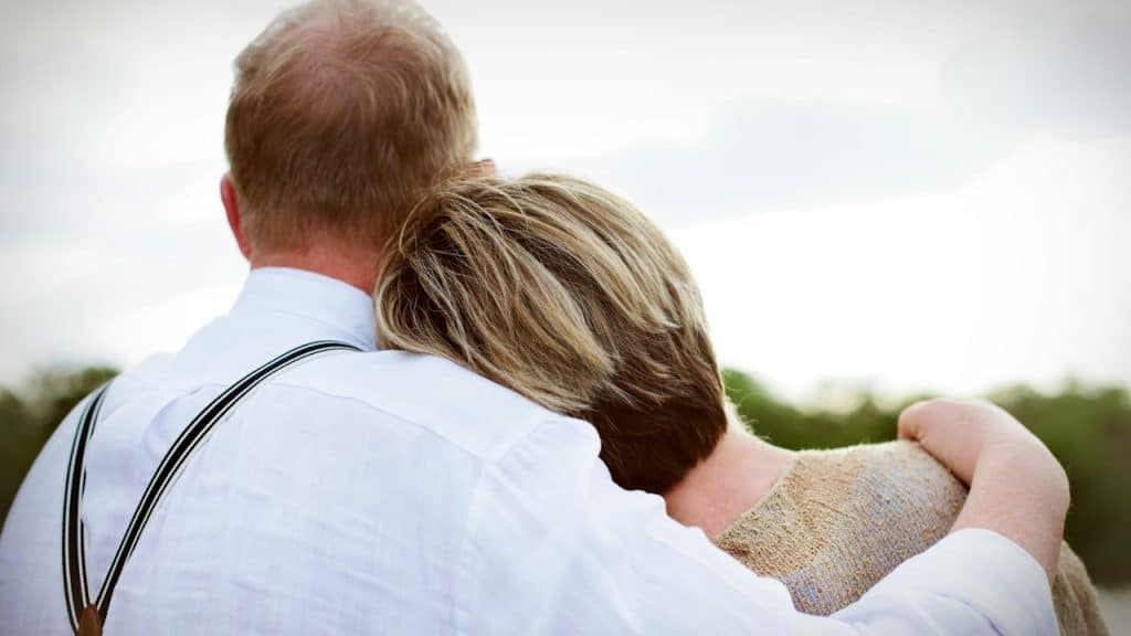 A close-up view from behind of an older couple embracing, with the woman resting her head on the man's shoulder.