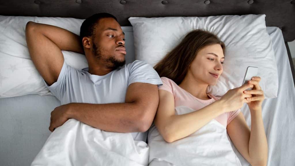 A man is lying in bed, looking suspiciously at the woman next to him who is smiling while looking at her phone.