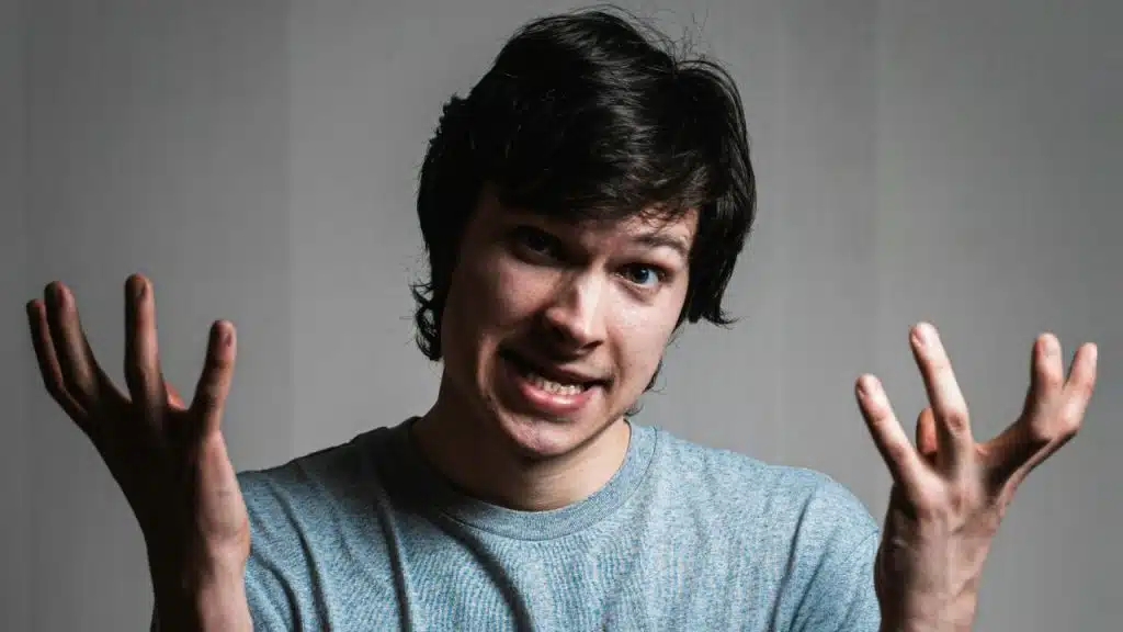 A young man with dark hair is shrugging, with his hands up and a skeptical expression.