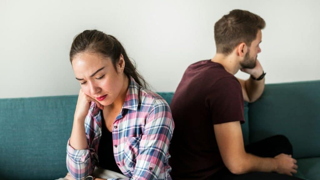 A woman and man sit back-to-back on a blue couch, both looking distressed.