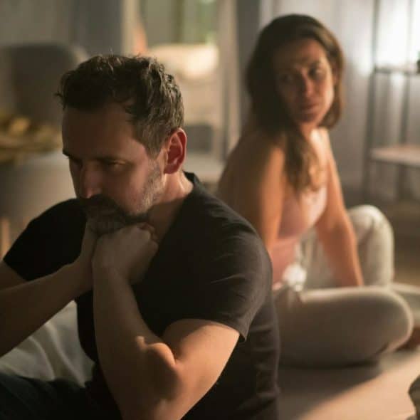 A man and woman sitting apart on a bed, looking upset in a dimly lit room.