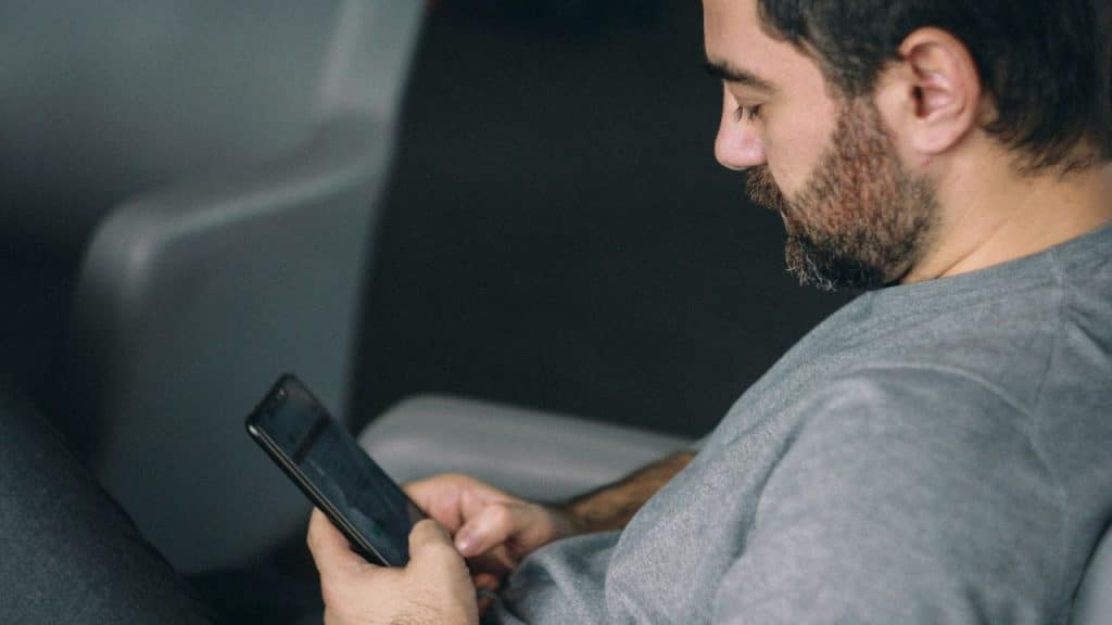 A bearded man in a gray shirt is concentrating on his mobile phone.