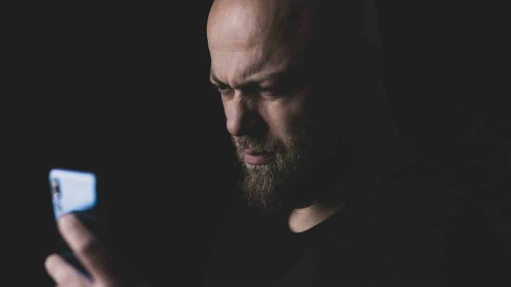 Bald, bearded man with a stern expression looking at a smartphone in the dark.