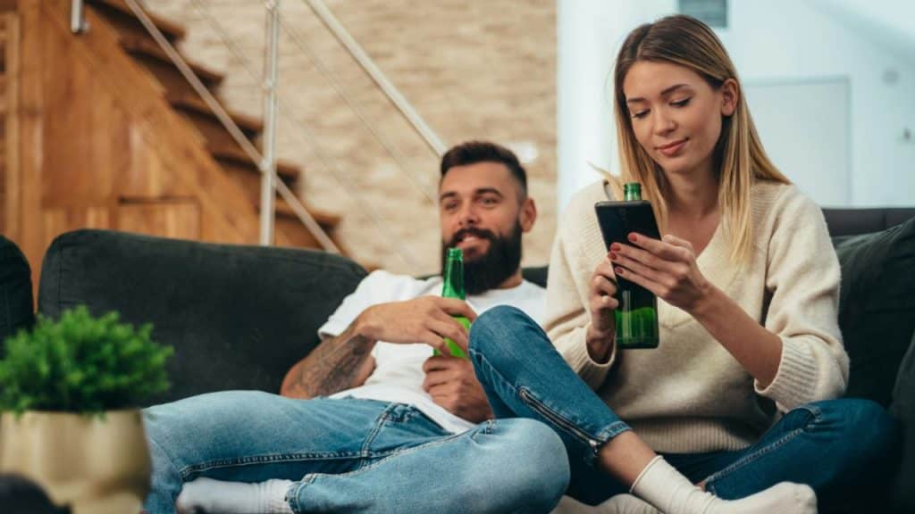 A woman focuses on her phone while a man smiles, holding a beer bottle.