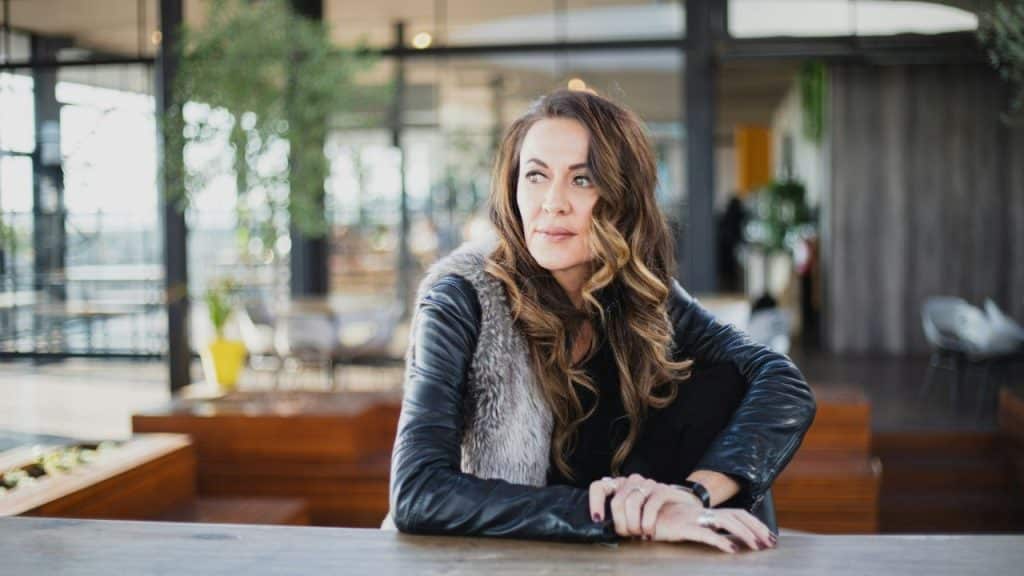 Woman in a black leather jacket and fur vest looking thoughtfully away at a bar or cafe.