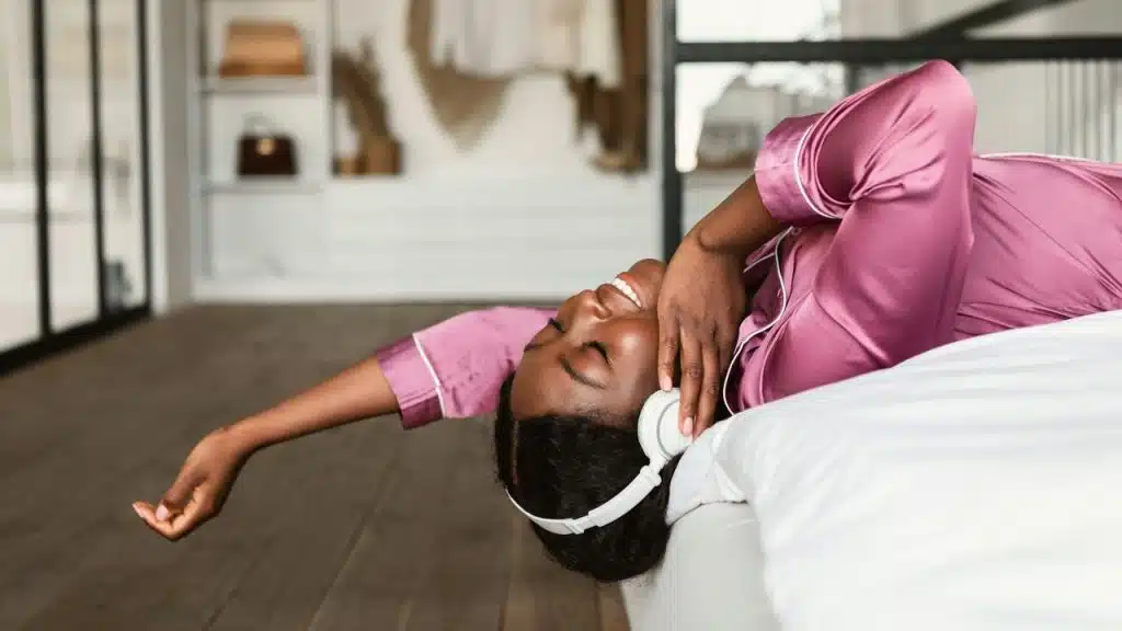 A woman in pink pajamas smiling and listening to music with headphones.