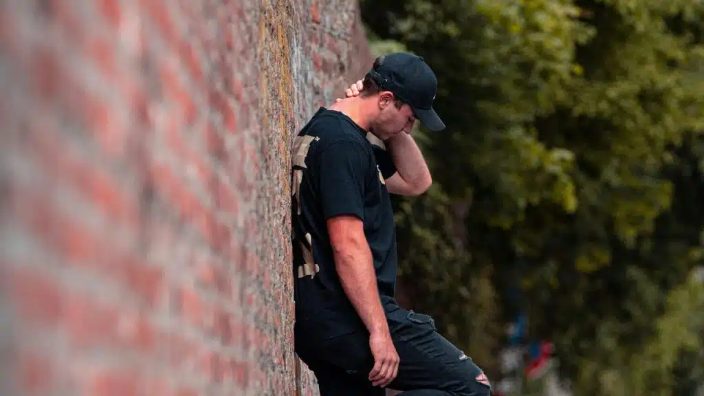 A man in a black shirt and cap leans against a brick wall, looking down.