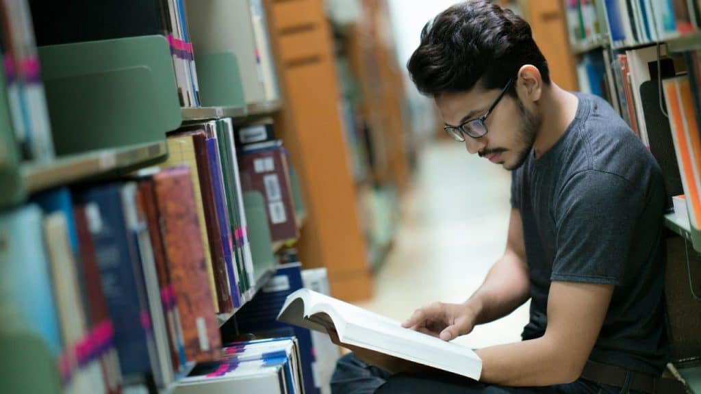 A young man with glasses sits on the floor between library shelves, reading a large book.