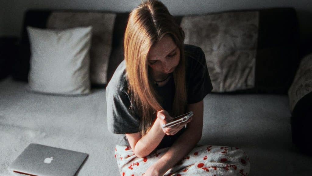 A woman sits on a couch in pajamas looking at her phone with a laptop beside her.