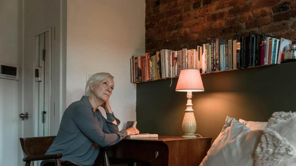 An older woman sitting at a desk resting her head on her hand near a lamp.