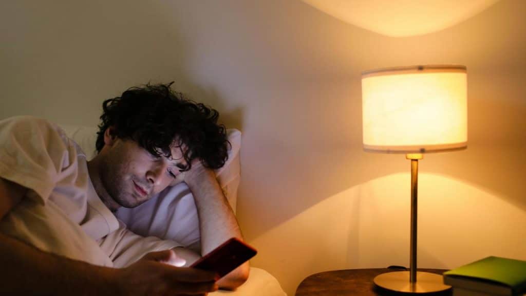 A man lying in bed at night looking at his phone beside a lit lamp.