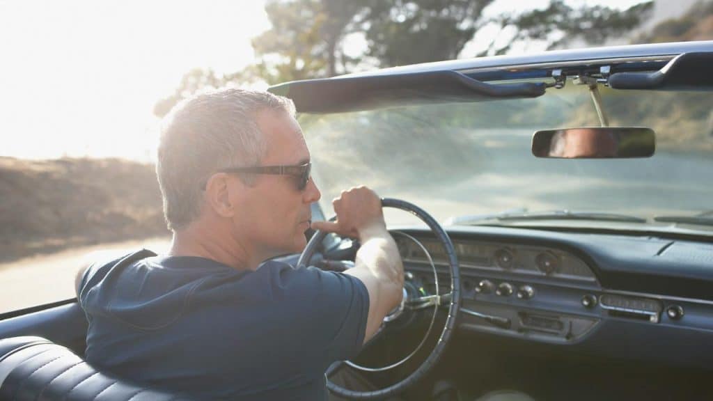 A man wearing sunglasses drives a vintage convertible on a sunny road.