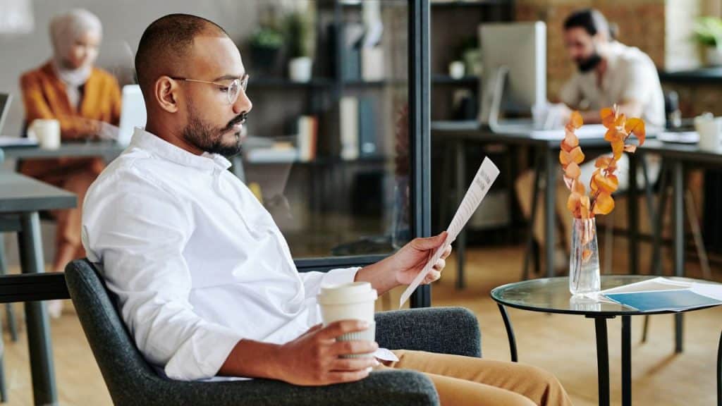 A man in glasses reading a document while holding a coffee cup in an office.