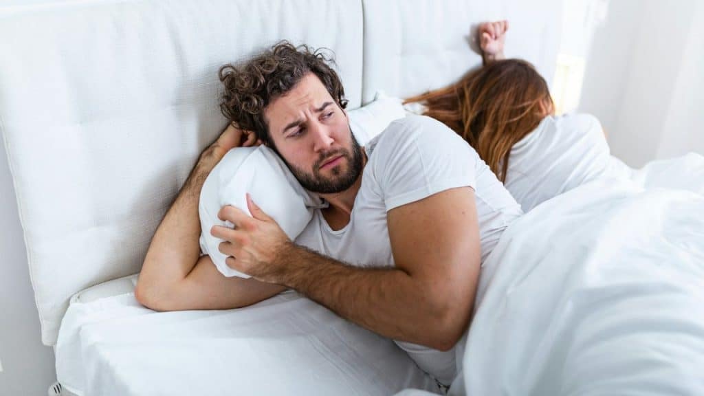 A sad man is lying in bed, awake, while a woman sleeps with her back to him.