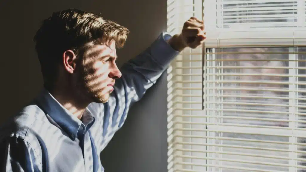 A serious man is looking out a window, holding up the blinds with a sunlit face.