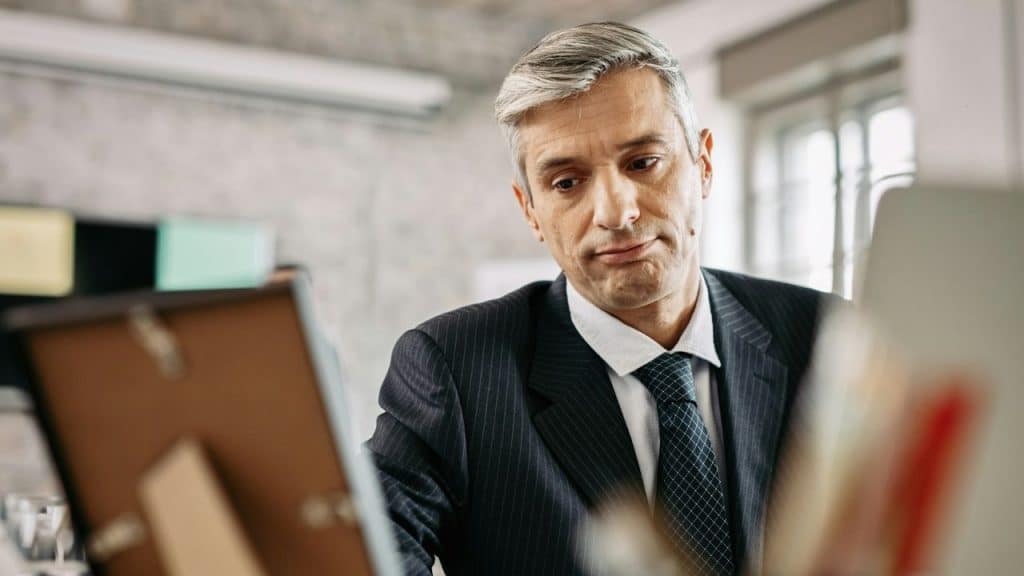 A middle-aged businessman in a pinstripe suit and tie looks down sadly at a photo in a frame on his desk.