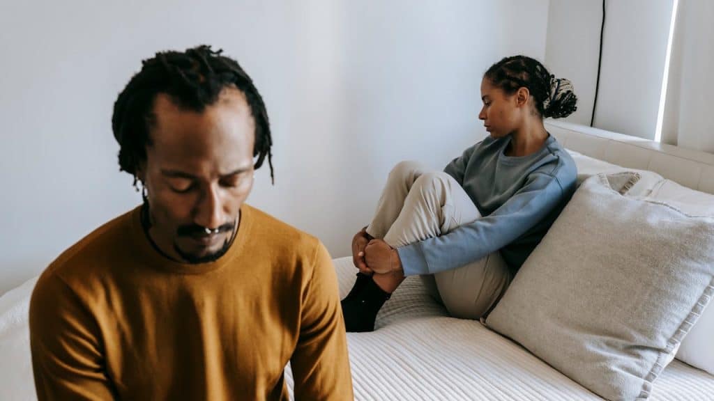 A sad man in a brown shirt is blurred in the foreground, while a distressed woman sits curled up on a bed in the background.