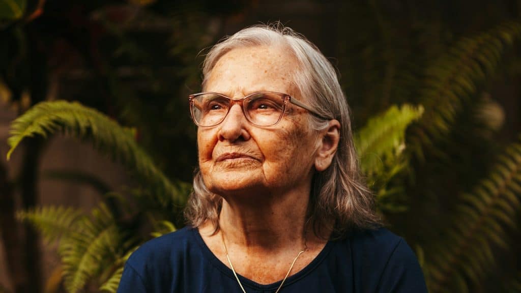 An older woman wearing glasses sitting in front of a plant.