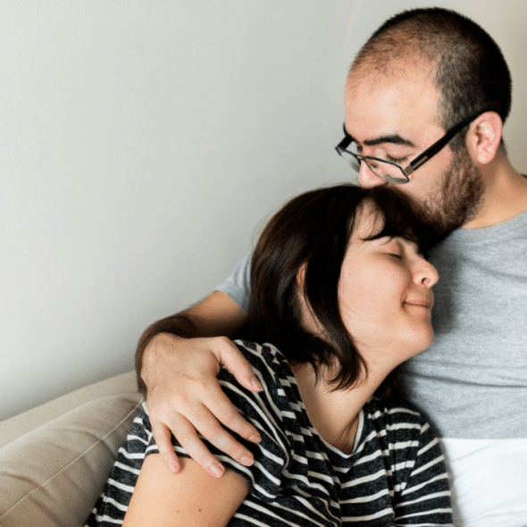 A man with glasses is hugging a woman on a couch and kissing her forehead.