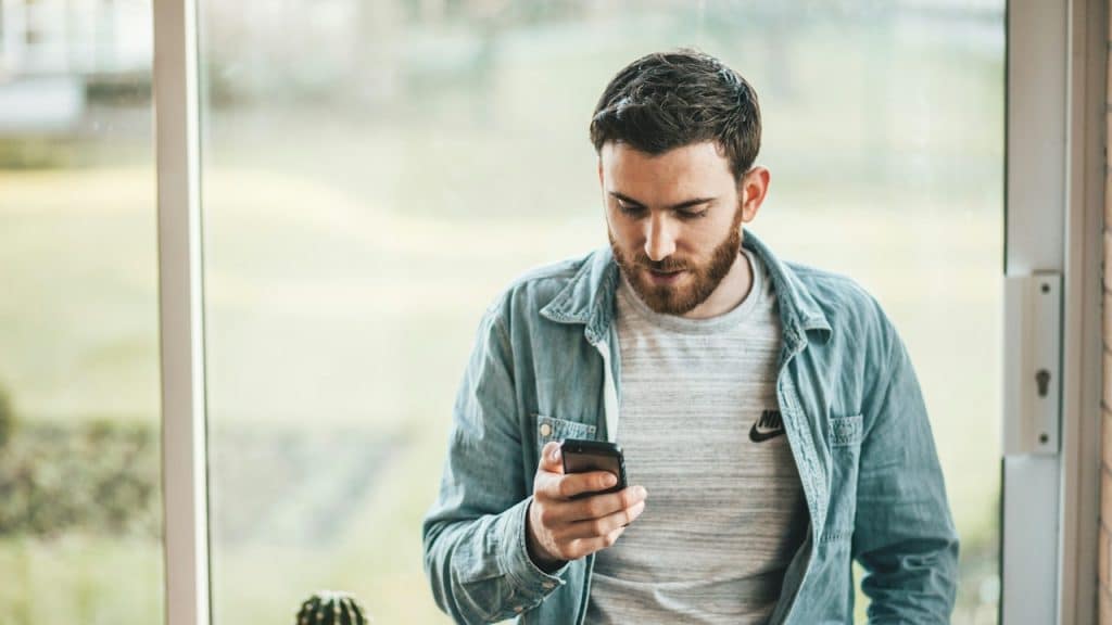 A casually dressed, bearded man in a denim shirt over a gray T-shirt stands by a bright window, intently looking down at his smartphone.