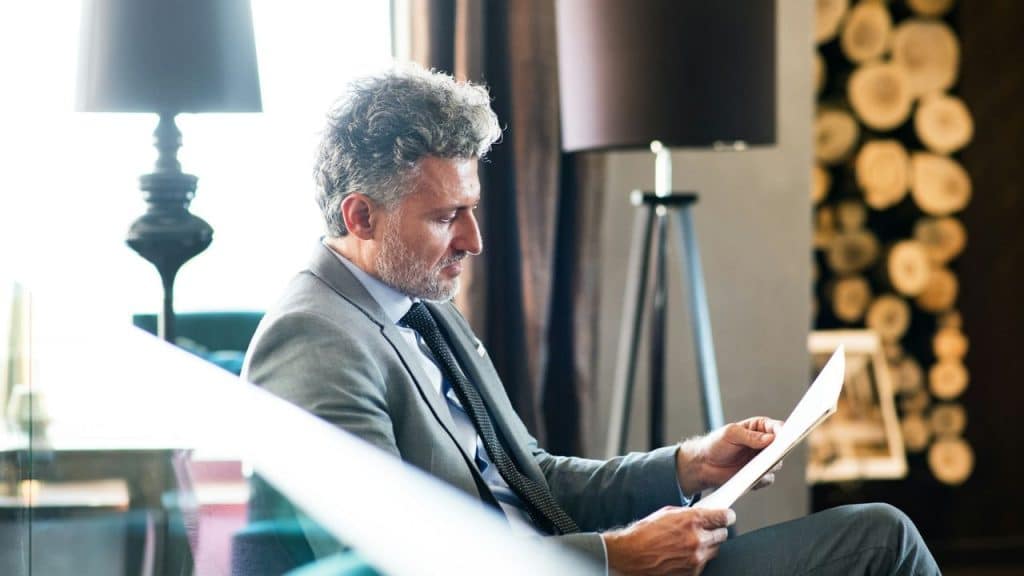 Man in a grey suit and tie reading a document indoors.