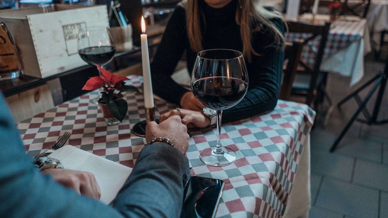A close-up of a couple holding hands across a checkered table, lit by a candle, with wine glasses in a restaurant.
