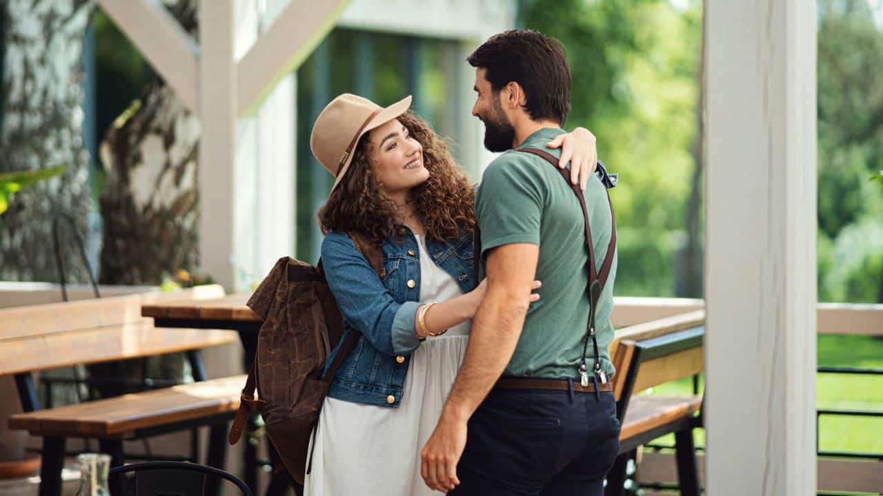 A smiling woman in a hat embraces a man goodbye at an outdoor cafe.