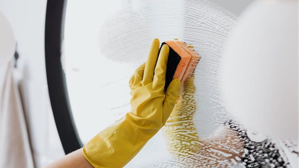A person wearing a yellow glove is cleaning a mirror with a soapy, orange and black sponge.