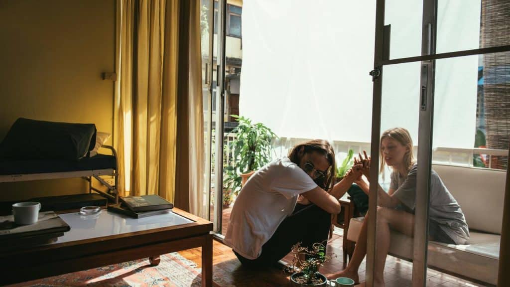 A man and woman sitting by a balcony door talking in a sunlit room.